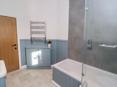 A bathroom with a bathtub and glass shower screen a wooden door a cabinet with flowers and a towel heater at West Burton Hemmel in Bamburgh
