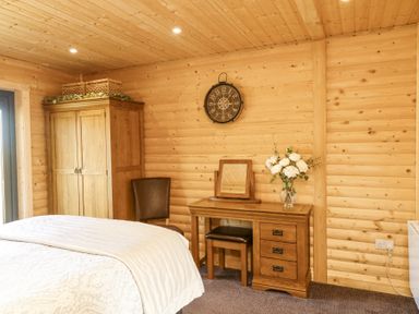 A bedroom with wooden walls and ceiling a bed a wooden wardrobe a chair a wooden dressing table with a mirror and flowers and a wall clock at Alpaca Lodge in Barkston