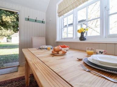 A wooden dining table with plates cups toast and a bowl of tomatoes next to a window with flowers at The Woodshed in Tendring near Weeley