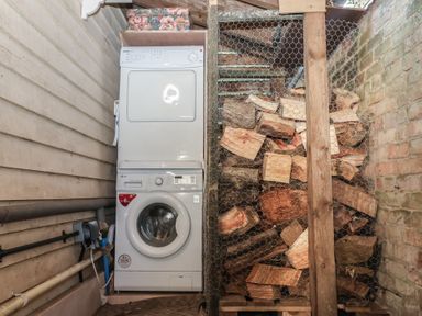 A laundry area with a stacked washer and dryer next to a storage area with firewood behind wire mesh at The Woodshed in Tendring near Weeley