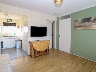 A room with a wall-mounted television above a wooden folding table and a view into a kitchen area at Chalet 469 in Newport Norfolk
