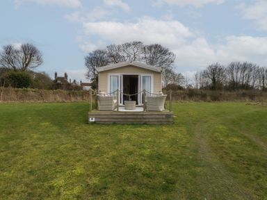 A small beige cabin with a wooden deck and outdoor chairs on a grassy lawn at Breathing Space in Happisburgh near Walcott