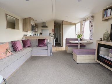 A living area with a grey sofa with purple pillows a dining nook with purple seating a kitchen area and a wall-mounted electric fireplace at Breathing Space in Happisburgh near Walcott