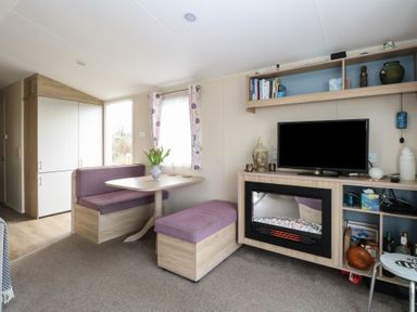 A small living area with a dining table, purple cushioned bench seats, a television above a fireplace, and shelves with books and decorative items at Breathing Space in Happisburgh near Walcott