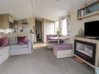 A living room with a sofa and purple cushions a dining area with a table and benches a fireplace and a television at Breathing Space in Happisburgh near Walcott