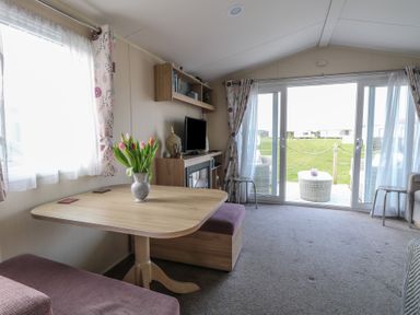 A living area with a wooden table and pink seating a television a bookshelf and sliding glass doors opening to a lawn at Breathing Space in Happisburgh near Walcott