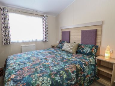 A bedroom with a bed covered in a floral patterned bedspread beside a window and wooden nightstand with a lamp at Breathing Space in Happisburgh near Walcott