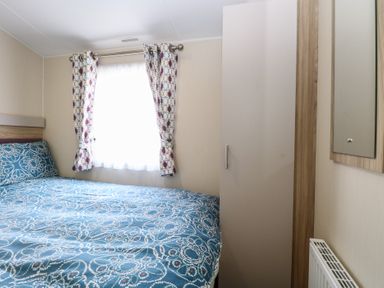 A bedroom with a bed covered in blue patterned bedding a window with patterned curtains a tall wardrobe a wall mirror and a radiator at Breathing Space Happisburgh near Walcott