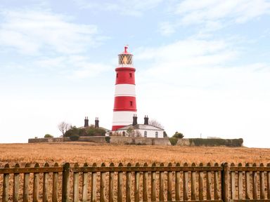 A red and white striped lighthouse behind a wooden fence with a field in front at Breathing Space Happisburgh near Walcott