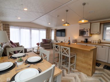 An open living space with a dining table set for four, a kitchen island with two stools, and a seating area with chairs and a sofa at Lodge 36 - Kingfisher Lodge in Wickhamford near Evesham