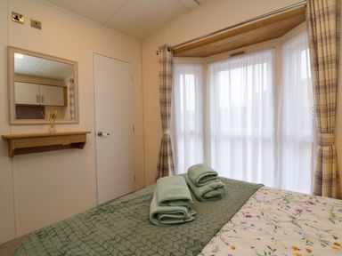 A bedroom with folded green towels on a bed next to a window with sheer curtains and plaid drapes at Lodge 36 - Kingfisher Lodge in Wickhamford near Evesham