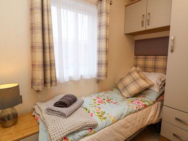 A single bed with floral bedding and plaid cushions next to a window with plaid curtains and a bedside table with a lamp at Lodge 36 - Kingfisher Lodge in Wickhamford near Evesham