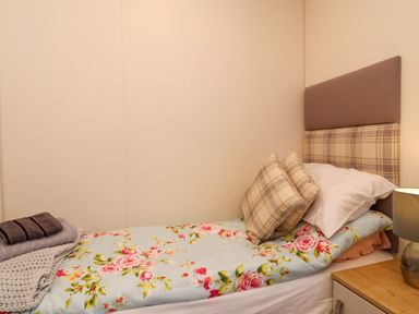 A single bed with floral bedding and plaid cushions next to a wooden nightstand with a lamp at Lodge 36 - Kingfisher Lodge in Wickhamford near Evesham