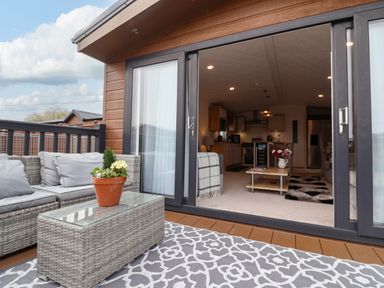An outdoor patio with grey wicker furniture and a potted plant overlooking an open sliding door into a living room and kitchen at Lodge 36 - Kingfisher Lodge in Wickhamford near Evesham