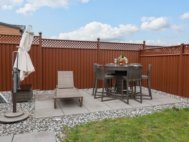 An outdoor patio with a wicker lounge chair a round wicker table with six chairs and a closed umbrella at Lodge 36 - Kingfisher Lodge in Wickhamford near Evesham