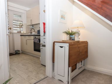 A view of a kitchen with white cabinets and a small wooden table with a lamp and plants under a staircase at Pie Cottage in Teignmouth