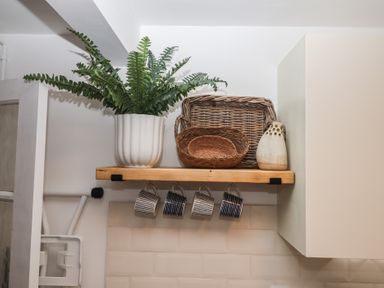 A wooden shelf with a white pot plant four hanging mugs two wicker trays and a small decorative vase at Pie Cottage in Teignmouth