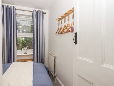 A small bedroom with a bed blue bedding white blanket a window with blue and white striped curtains and wooden hangers on wall at Pie Cottage in Teignmouth