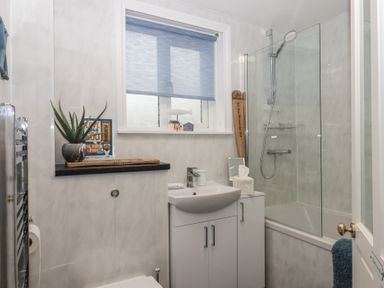 A small bathroom with a sink cabinet bathtub with glass shower screen and blue window blind at Pie Cottage in Teignmouth