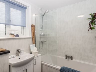 A bathroom with a sink, bathtub with shower, window with blind, and decorative items at Pie Cottage in Teignmouth
