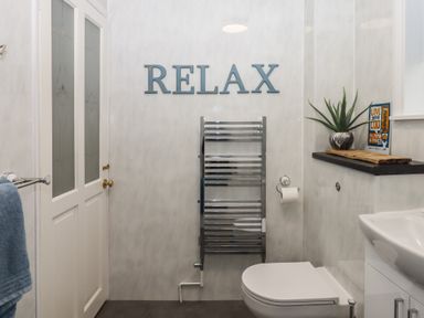 A bathroom with a toilet sink towel rail and a plant on a shelf with relax sign on wall at Pie Cottage in Teignmouth