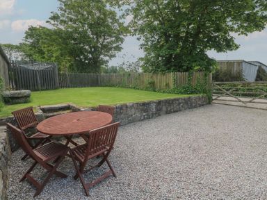 An outdoor area with a wooden table and four chairs on gravel next to a grass lawn with a picket fence and trees at Sea View Loft in Rosudgeon