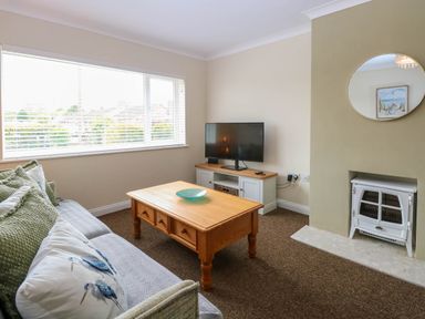 A living room with a sofa with cushions a wooden coffee table a flat screen TV on a cabinet a fireplace and a round mirror at Little Lodge in Dersingham