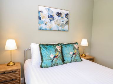 A bedroom with a bed featuring two bird-themed pillows two bedside tables with lamps and floral artwork above the headboard at Little Lodge in Dersingham