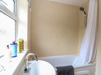 A bathroom with a bathtub shower combination with a white shower curtain and small yellow tiled walls with toiletries on the window ledge at Little Lodge in Dersingham