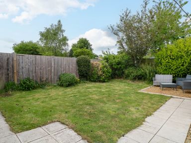 A backyard with grass lawn, a wooden fence, bushes, trees, and outdoor seating on gravel at Little Lodge in Dersingham