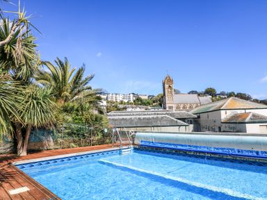 An outdoor swimming pool with palm trees and buildings including a church tower in the background at Delmonte Lodge in Torquay