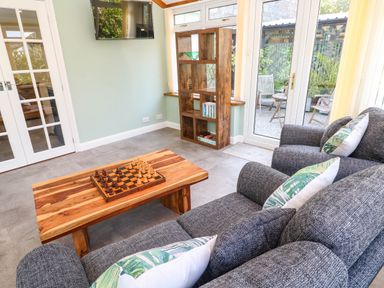 A sitting area with gray armchairs a wooden coffee table with a chessboard a wooden bookshelf and glass doors leading outside at Little Acorns in Gweek