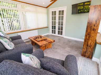 A sitting room with two gray armchairs a wooden coffee table with a chess set and a wall-mounted television at Little Acorns in Gweek