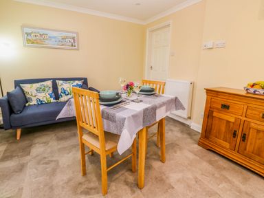 A dining area with a wooden table and two chairs set for two with plates and wine glasses next to a blue couch with floral cushions and a wooden sideboard at Little Acorns in Gweek