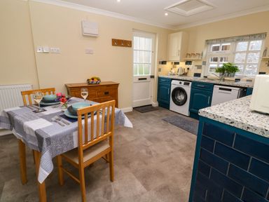A kitchen with a dining table set for two wooden chairs a washing machine blue cabinets and a wooden sideboard at Little Acorns in Gweek