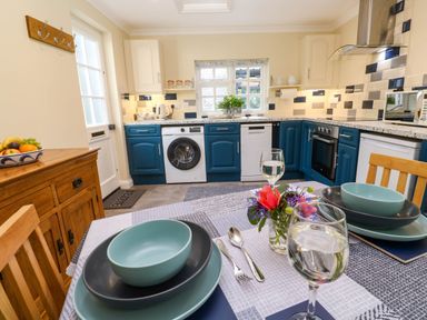 A kitchen with blue and white cabinets a dining table set with plates bowls glasses and flowers at Little Acorns in Gweek