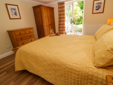A bedroom with a wooden dresser and wardrobe a bed with yellow bedding and a tray with glasses and strawberries by glass doors opening to a garden at Little Acorns in Gweek