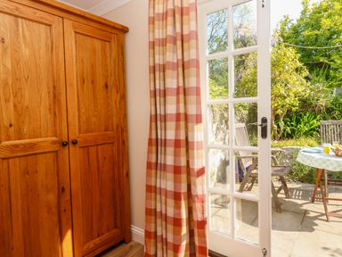 A wooden wardrobe next to glass doors with checkered curtains opening to a garden patio with chairs and a table at Little Acorns in Gweek