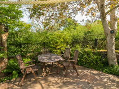 An outdoor patio area with a wooden table and four wooden chairs surrounded by trees and greenery at Little Acorns in Gweek