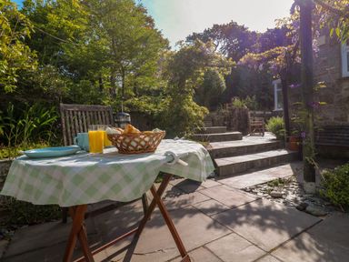 An outdoor patio with a table covered by a checkered tablecloth set with glasses of orange juice and a basket of bread near stone steps and greenery at Little Acorns in Gweek
