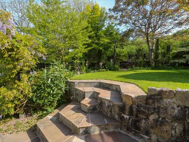 A garden with stone steps leading to a grassy area surrounded by trees and plants at Little Acorns in Gweek