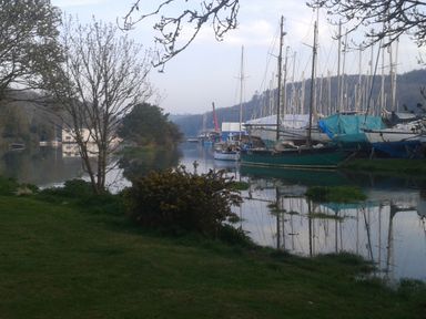 A riverside view with anchored sailboats and a grassy bank with trees and shrubs at Little Acorns in Gweek