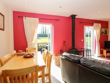 A dining area with wooden table and chairs next to a living room with a black leather sofa and a wood stove in front of red walls at Swallow Barn in Holton near Halesworth