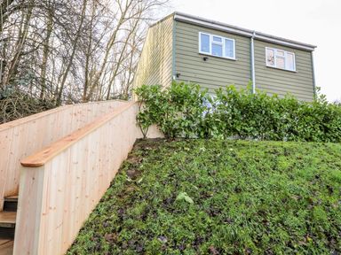 An exterior view of a green house with white windows behind a hedge and wooden railing on a grassy slope at Arnside House in South Lakeland Leisure Village