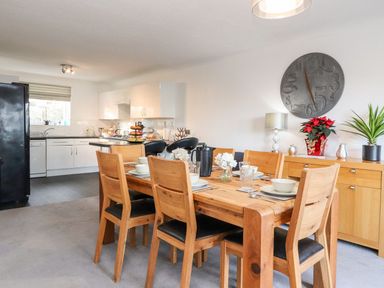 A dining area with a wooden table set with dishes and glasses next to a kitchen with white cabinets and black appliances at Arnside House in South Lakeland Leisure Village