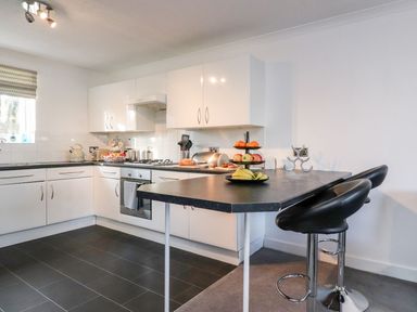 A kitchen with white cabinets black countertops a stove and a breakfast bar with two black stools at Arnside House South Lakeland Leisure Village