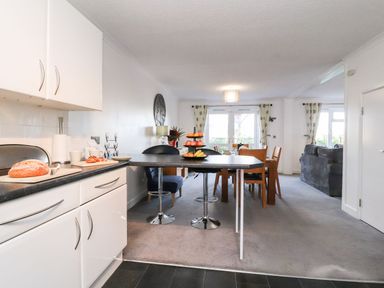 A kitchen with a countertop and stools opening to a dining area with a table and chairs and a living room with sofas at Arnside House in South Lakeland Leisure Village