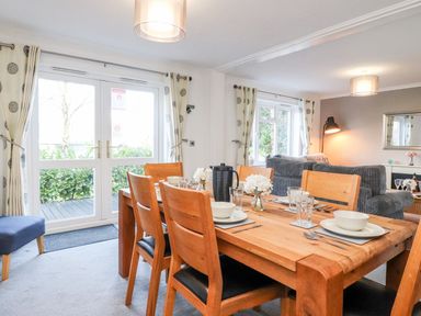 A dining area with a wooden table set for four and adjacent living room with sofas and lamps at Arnside House South Lakeland Leisure Village