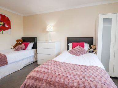 A bedroom with two single beds pink pillows and teddy bears a white chest of drawers with a lamp and a white wardrobe at Arnside House in South Lakeland Leisure Village