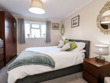 A bedroom with a bed with pillows and blanket below a window with curtains and a bedside table with lamp next to a wardrobe at Arnside House in South Lakeland Leisure Village